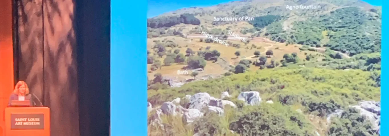 an auditorium stage with a slide of Mt. Lykion and Voyatzis speaking at a podium