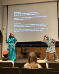 two students in masks perform a scene from a Roman comedy with Latin text on the screen above them