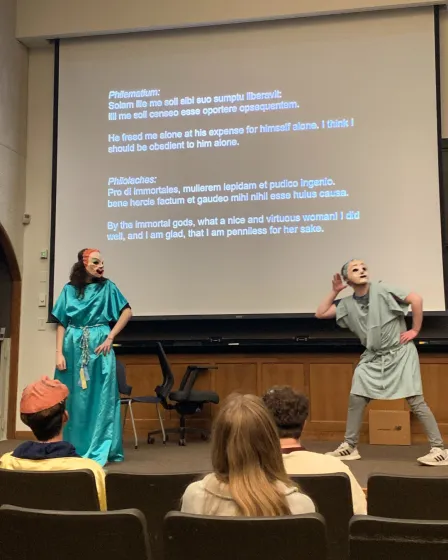two students in masks perform a scene from a Roman comedy with Latin text on the screen above them